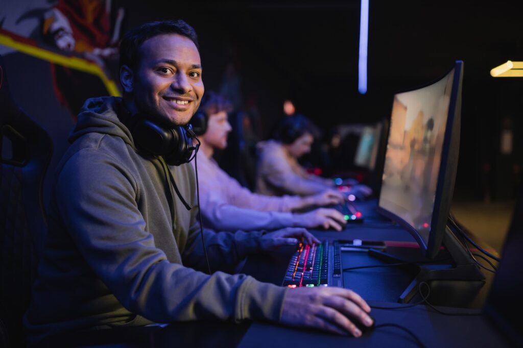 Smiling indian gamer looking at camera near computer in cyber club